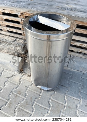 A tall gray metal urn with a cylindrical frame stands on gray textured paving slabs near a wooden bench, close-up, side view. Special place for garbage. Let's save nature.