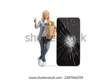 Woman with a grocery bag leaning on a big mobile phone with broken screen and showing thumbs up isolated on white background