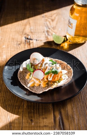 Overhead of a crispy fish taco on dark plate with sauce drizzle and beer with a sliced lime in the background.