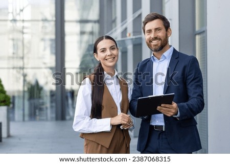 Two happy male and female business colleagues in suits standing outside near office, discussing important work issues, comparing papers, preparing for presentation, looking at camera and smiling. Royalty-Free Stock Photo #2387068315