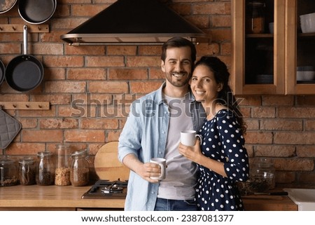 Family portrait happy young couple drinking coffee or tea in morning, standing in cozy kitchen at home, smiling wife and husband hugging, looking at camera, holding mugs, starting new day together