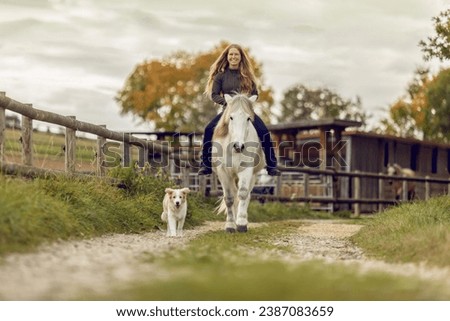 Horse and dog concept: A young female equestrian and her dog on her icelandic horse in front of a rural farm landscape Royalty-Free Stock Photo #2387083659