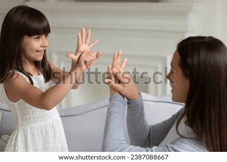 Smiling little girl and young mother speaking sign language, showing gestures, sitting on cozy sofa at home, happy preschool daughter and mum communicating, having fun, playing game with hands Royalty-Free Stock Photo #2387088667