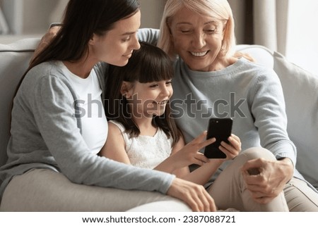 Happy little girl with mother and grandmother using smartphone together close up, three generations of women sitting on sofa, having fun with phone together, watching video or shopping online