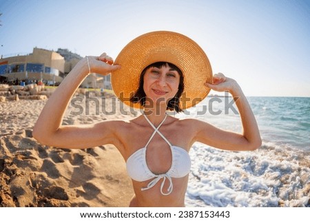 Portrait of a beautiful young woman in a straw hat on the seashore. Cheerful young woman smiling on the beach during summer vacation. Happy girl with black hair enjoying the sun.