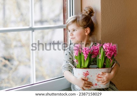 Little toddler girl sitting by window with blossoming pink hyacinth flowers. Happy child, indoors. Mother's day, valentine's day or birthday and spring concept. Royalty-Free Stock Photo #2387158029