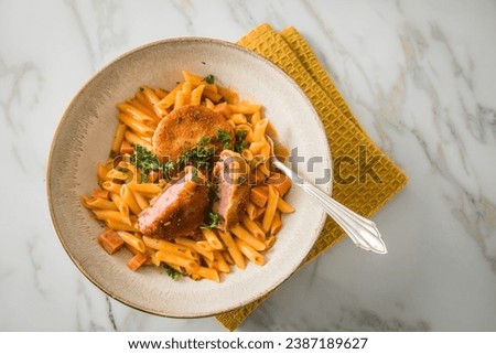 Jägerschnitzel escalope chasseur a East German DDR GDR specialty of breaded fried sausage slice, tomato sauce, Macaroni Penne pasta, parsley in bowl, cutlery, napkin, marble background Royalty-Free Stock Photo #2387189627