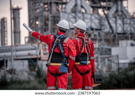 Back side view engineer survey team wear uniform and helmet stand workplace checking blueprint project and engineer box  inspection work construction site with oil refinery background. Royalty-Free Stock Photo #2387197063