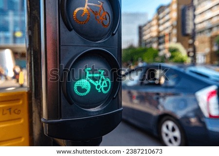 A green cycle lane traffic  signal at a junction in a busy London city street with a car passing in the background.
