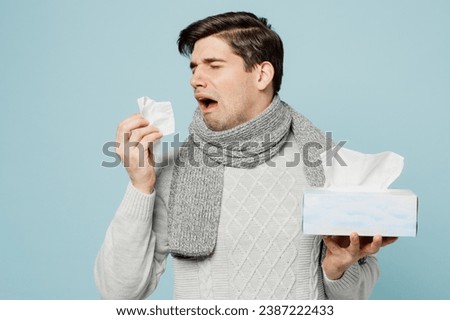 Young ill sick man wear gray sweater scarf sneezing hold box of paper napkins isolated on plain blue background studio portrait. Healthy lifestyle disease virus treatment cold season recovery concept