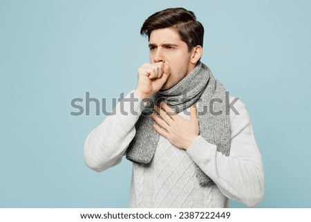 Young ill sick man wear gray sweater scarf cough sneeze cover mouth with hand isolated on plain blue background studio portrait. Healthy lifestyle disease virus treatment cold season recovery concept Royalty-Free Stock Photo #2387222449