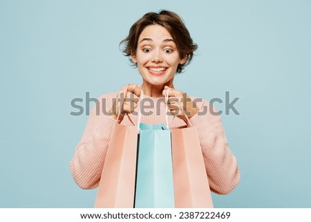 Young surprised woman wear beige knitted sweater casual clothes hold in hand open paper package bags after shopping isolated on plain light pastel blue background. Black Friday sale buy day concept