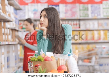 Bored young woman leaning on the shopping cart at the grocery store, she is looking around and feeling tired
