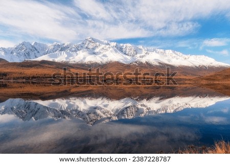 Beautiful autumn mountain lake and mountains. Reflection of snowy peaks and red coniferous taiga on the surface of a mountain lake. Russia. Altai Republic. Panoramic view. 