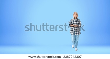 Full length portrait of cheerful young woman college or university student with blond hair wearing casual clothes and holding folder. Blue copy space background