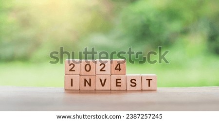  wooden blocks spelling ‘2024 INVEST’ on a rustic table, set against a blurred backdrop of lush greenery, concept of financial trends, investment opportunities.