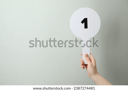 Woman holding auction paddle with number 1 on light grey background, closeup. Space for text