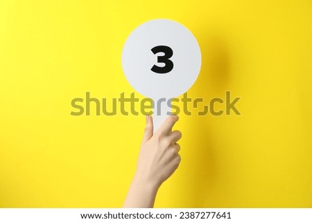 Woman holding auction paddle with number 3 on yellow background, closeup Royalty-Free Stock Photo #2387277641