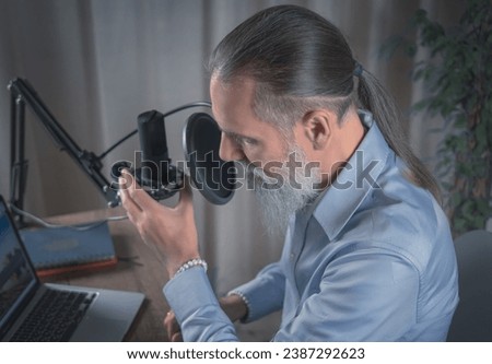 A male Presenter with a beard prepares to broadcast his audio podcast using a microphone and laptop in his home studio. Close-up