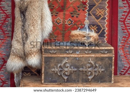 Traditional Asian wooden chests with carved ornaments, animal skins and a fur men's national hat against the background of the felt walls of a yurt - the home of nomads. Close-up Royalty-Free Stock Photo #2387294649