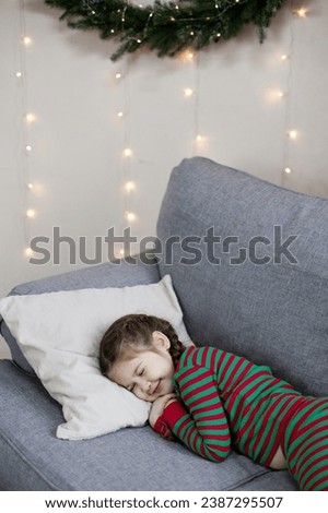 Preschool caucasian girl in Christmas pyjamas slepping on a grey sofa near Christmas tree with decorations