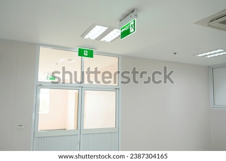 Selective fire exit sign on white ceiling and glass door.Green fire escape sign hang on the ceiling in the office.Fire fighting equipment concept.