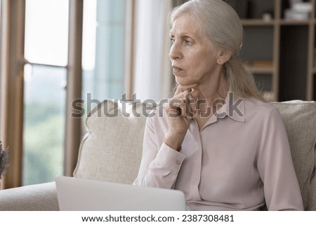 Serious senior pensioner woman looks thoughtful while working on laptop sit on sofa at home staring into distance, faced up with difficulties, modern tech usage problem, lack of understating need help Royalty-Free Stock Photo #2387308481