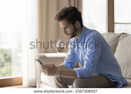 Serious millennial bearded man using cellphone seated on sofa at home. Single guy make call, check messages, answer on sms, chatting on-line using mobile application. Modern tech, remote communication Royalty-Free Stock Photo #2387308507