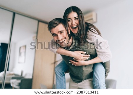 Happy man giving piggyback ride to laughing girlfriend in apartment Royalty-Free Stock Photo #2387313411