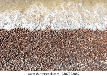 A white foaming sea wave washes the brown-red pebbles of the coast. Top view photo.