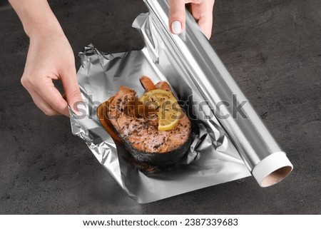 Woman wrapping tasty salmon with lemon and spices in aluminum foil at grey textured table, closeup Royalty-Free Stock Photo #2387339683