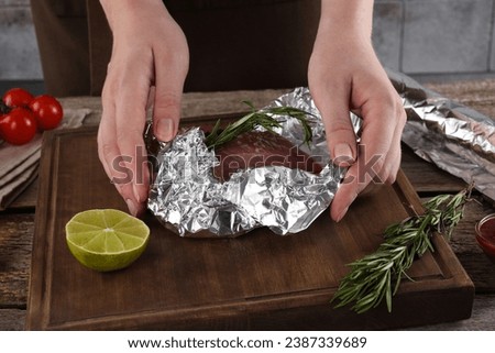 Woman wrapping meat in aluminum foil at wooden table, closeup Royalty-Free Stock Photo #2387339689