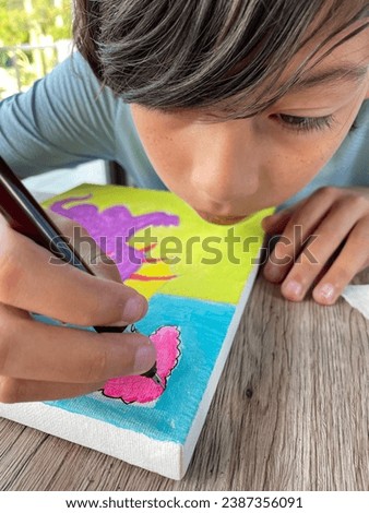 Photo of a young handsome good looking make kid boy artist who is painting a little purple or violet cute adorable cartoony dragon on a canvas looking like a master piece as he is talented and skilled