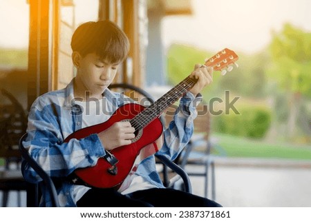 Asian cute boy sits infront of glassdoor of balcony and playing acoustic guitar, soft and selective focus, sunset light edited background.