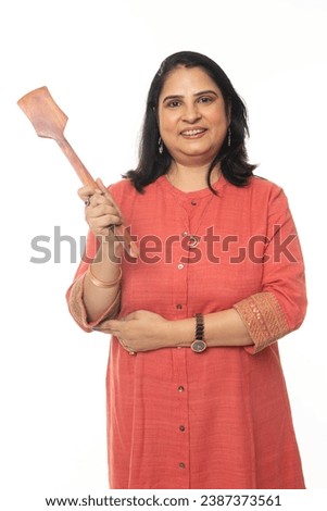 Indian happy woman holding wooden spatula. House wife showing kitchen items and home appliances products for cooking food. while standing isolated over white background Royalty-Free Stock Photo #2387373561
