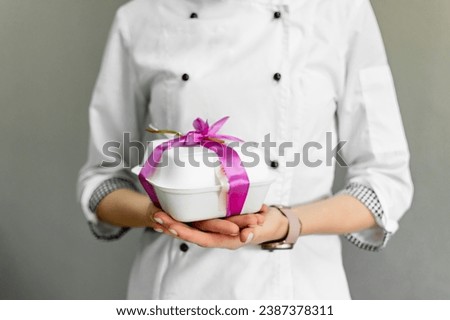 a pastry chef in a white uniform holds in his hands a small box with a cake tied with a pink ribbon