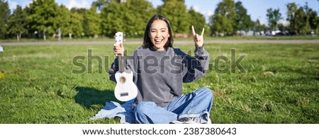 Positive korean girl sits in park, shows ukulele and rock on gesture, learns how to play musical instrument outdoors. Royalty-Free Stock Photo #2387380643