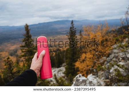 Pink thermos in the hand of a man on the background of autumn forest, concept of drinking tea in the mountains in nature. Trekking in the mountains. High quality photo