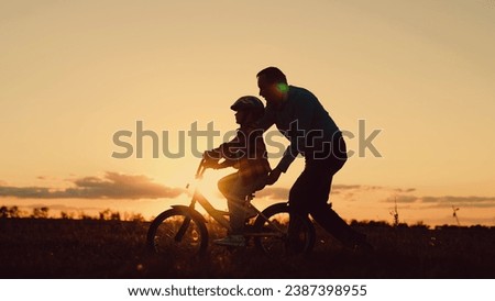 Father helps his daughter ride bike. Father teaches child wearing safety helmet to ride bicycle in park. Child rides bicycle. Kid, dad play together, sunset. Child dream learns to ride bicycle. Family Royalty-Free Stock Photo #2387398955