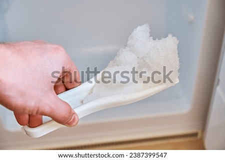 A man holds a shovel with ice on the background of a freezer. Defrosting the freezer. Royalty-Free Stock Photo #2387399547