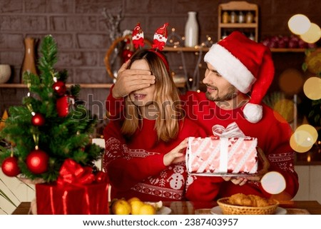 happy lovers a man and a girl in red sweaters in the kitchen with a Christmas tree give each other gifts closing their eyes at home and celebrate New Year or Christmas rejoicing and smiling Royalty-Free Stock Photo #2387403957