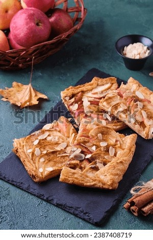 Sliced open apple pie on puff pastry with apple slices, sprinkled with almonds, on a black slate board on a green concrete background. Recipes apple pie, puff pastry