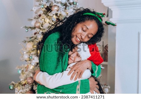 African American woman with curly hair and brunette daughter pose hugging and wearing Christmas hats against tree decorations
