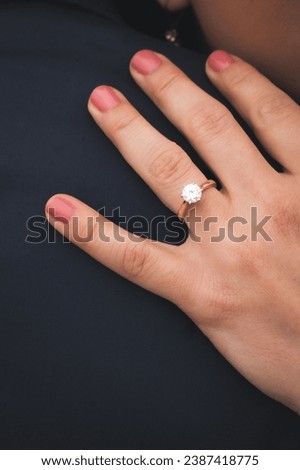 A close-up of a hand with a pink manicured nail resting on a dark fabric, adorned with a stunning gold engagement ring featuring a sparkling diamond Royalty-Free Stock Photo #2387418775
