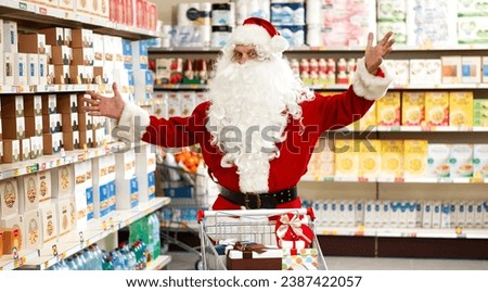 Happy Santa Claus doing grocery shopping at the supermarket, he is smiling with raised arms and looking at camera Royalty-Free Stock Photo #2387422057