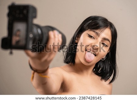 Girl holding a camera, taking a selfie showing her tongue on a pastel background.