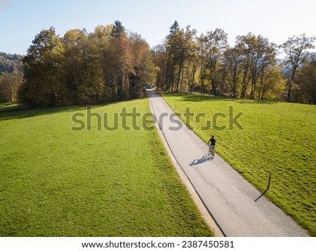 Cyclist riding along a quiet countryside road near green meadows and colorful woods on a sunny autumn day, aerial shot. Cycling tourism concept.