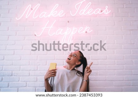 Happy woman with smartphone in hand near white brick wall