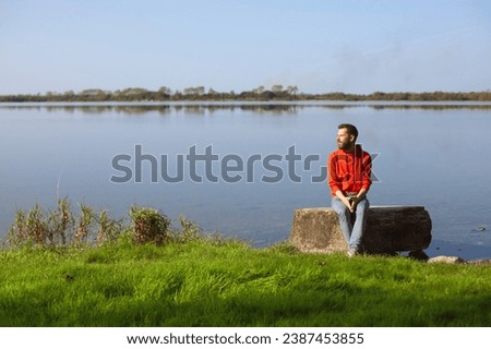 Discovering Ireland concept. Portrait of a young handsome man in red hoodie against the backdrop Lough Ennell in Belvedere House near Mullingar, County Westmeath. Outdoor shot Royalty-Free Stock Photo #2387453855