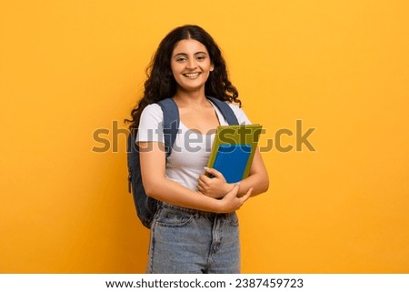 Education Concept. Positive smiling indian woman student holding notebooks looking at camera, copy space for advertisement. Woman wearing backpack, yellow studio background Royalty-Free Stock Photo #2387459723